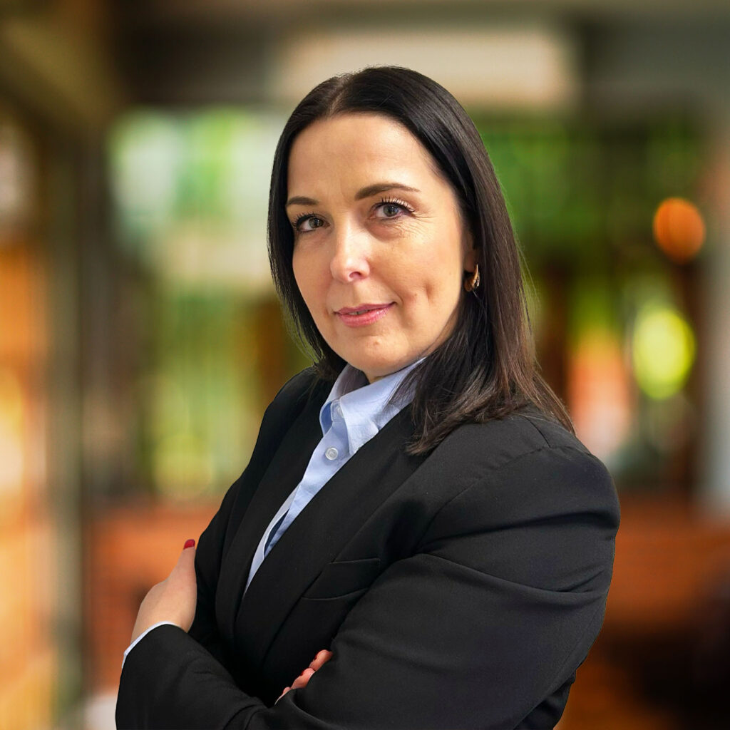 A professional headshot of a woman with dark hair, wearing a black blazer and a white shirt. She has a confident expression with her arms crossed and is facing the camera. The background is out of focus with warm hues, likely indoors.