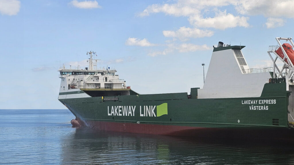 A large green and white cargo ship named "LAKEWAY EXPRESS VÄSTERÅS" is docked in calm blue waters under a partly cloudy sky. The ship's hull is prominently visible with the name "LAKEWAY LINK" painted on the side in large white letters. The vessel's structure features a bridge deck and various other operational decks towards the stern. There is also a white loading ramp folded onto the stern deck, indicating that this ship is likely a Ro-Ro (roll-on/roll-off) vessel designed for carrying wheeled cargo.