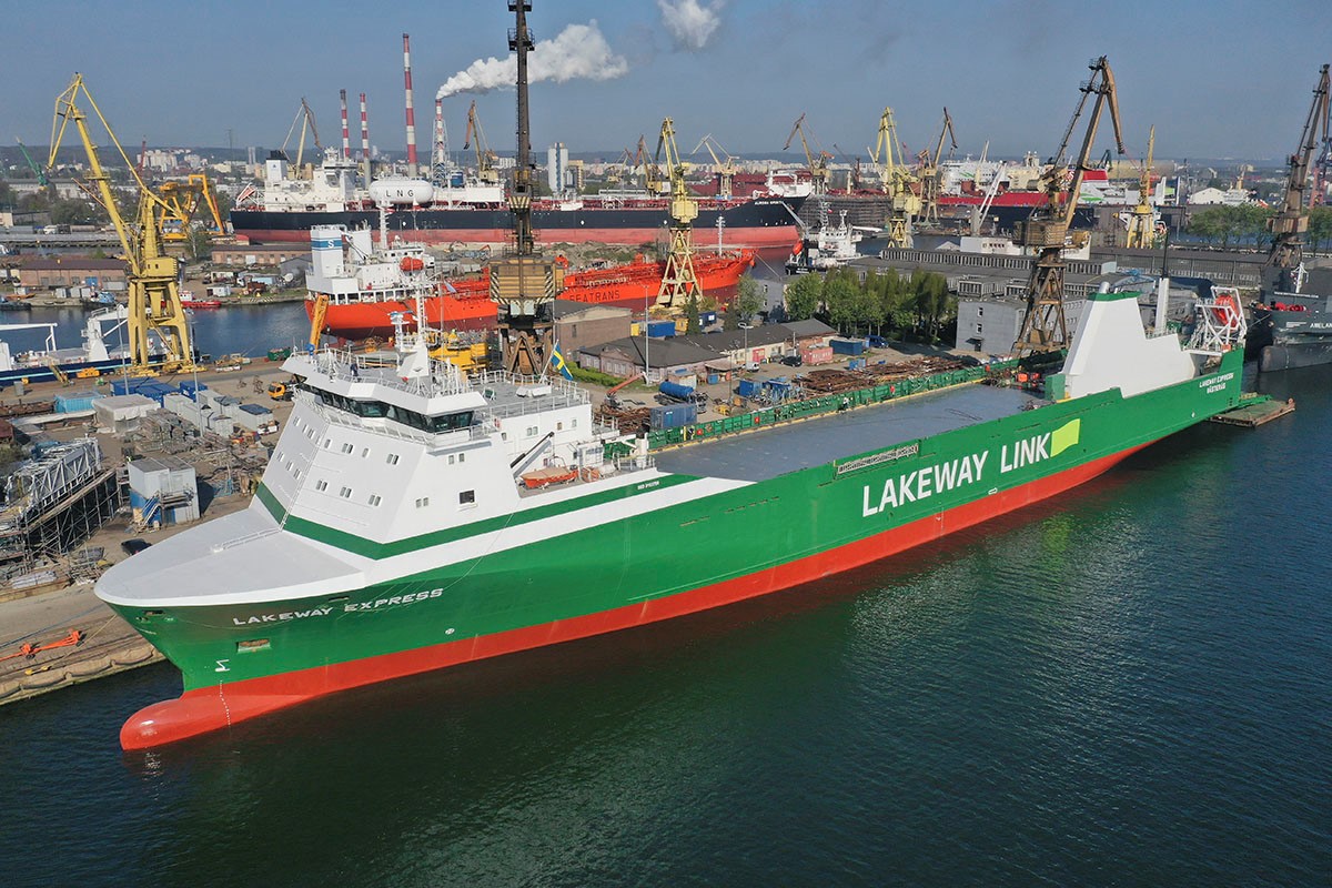 A large green and white cargo ship named "Lakeway Express" is docked at a busy industrial shipyard. The ship features the branding "Lakeway Link" along its side. The surrounding area is filled with cranes, containers, and other ships, including a prominent LNG tanker and another large vessel painted orange. The shipyard is active with various industrial structures and equipment visible. Smoke rises from chimneys in the background, indicating industrial activity.