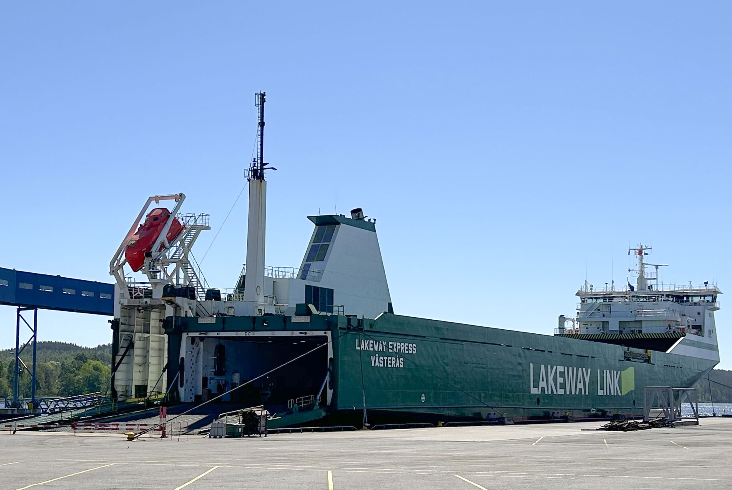 A large green and white cargo ship named "Lakeway Express Västerås" is docked at a port. The ship features the branding "Lakeway Link" prominently on its side. The ship has a red lifeboat mounted on its upper deck, and its ramp is lowered, indicating it might be in the process of loading or unloading cargo. The sky is clear and blue, and there are trees and hills visible in the background, suggesting a calm and pleasant day at the port. The ground around the dock is empty, with a few scattered objects and equipment.