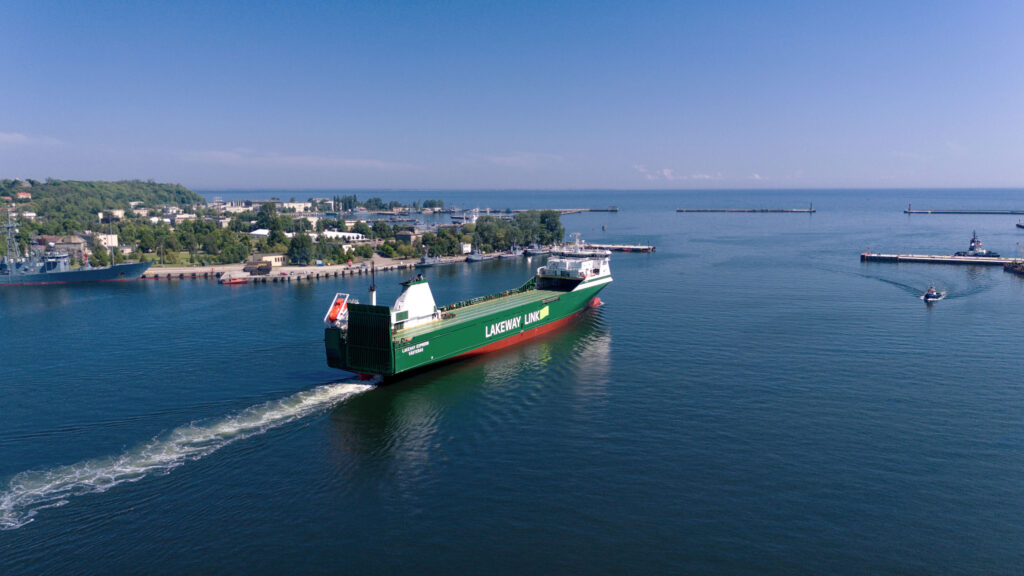 A large green cargo ship named "Lakeway Link" is sailing away from a harbor on a clear, sunny day. The ship is prominently marked with the name "Lakeway Express" and is surrounded by calm blue waters. The harbor area has various buildings, trees, and a docked navy ship. Small boats are visible in the water, and breakwaters extend into the lake, defining the harbor's entrance. The sky is clear with a few scattered clouds, and the overall scene conveys a sense of serene maritime activity.