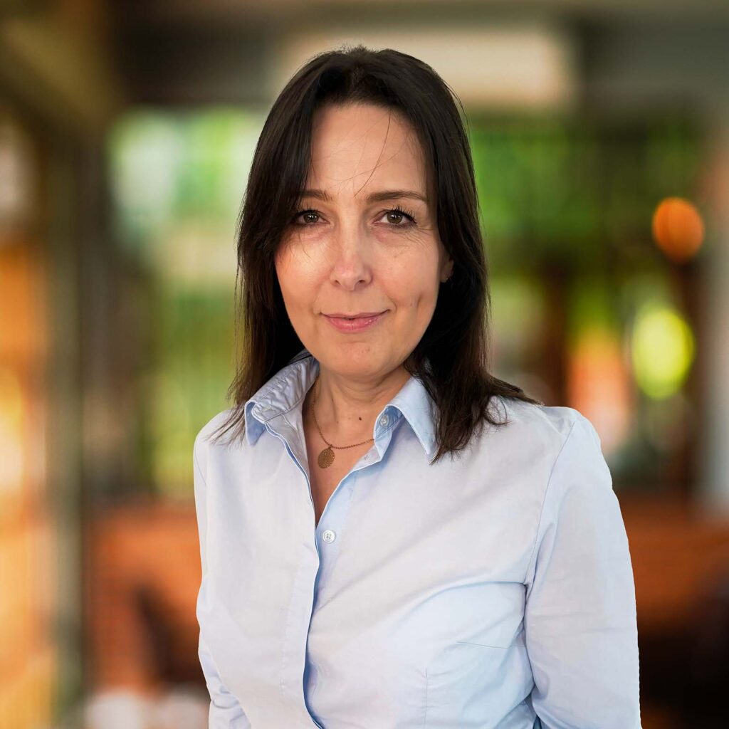 The image shows a woman with dark brown hair and a light complexion, wearing a light blue button-up shirt. She has a slight smile and is looking directly at the camera. The background appears to be blurred, suggesting an indoor setting with warm lighting, possibly a cafe or a similar space with wooden accents and large windows. The overall atmosphere is warm and inviting.