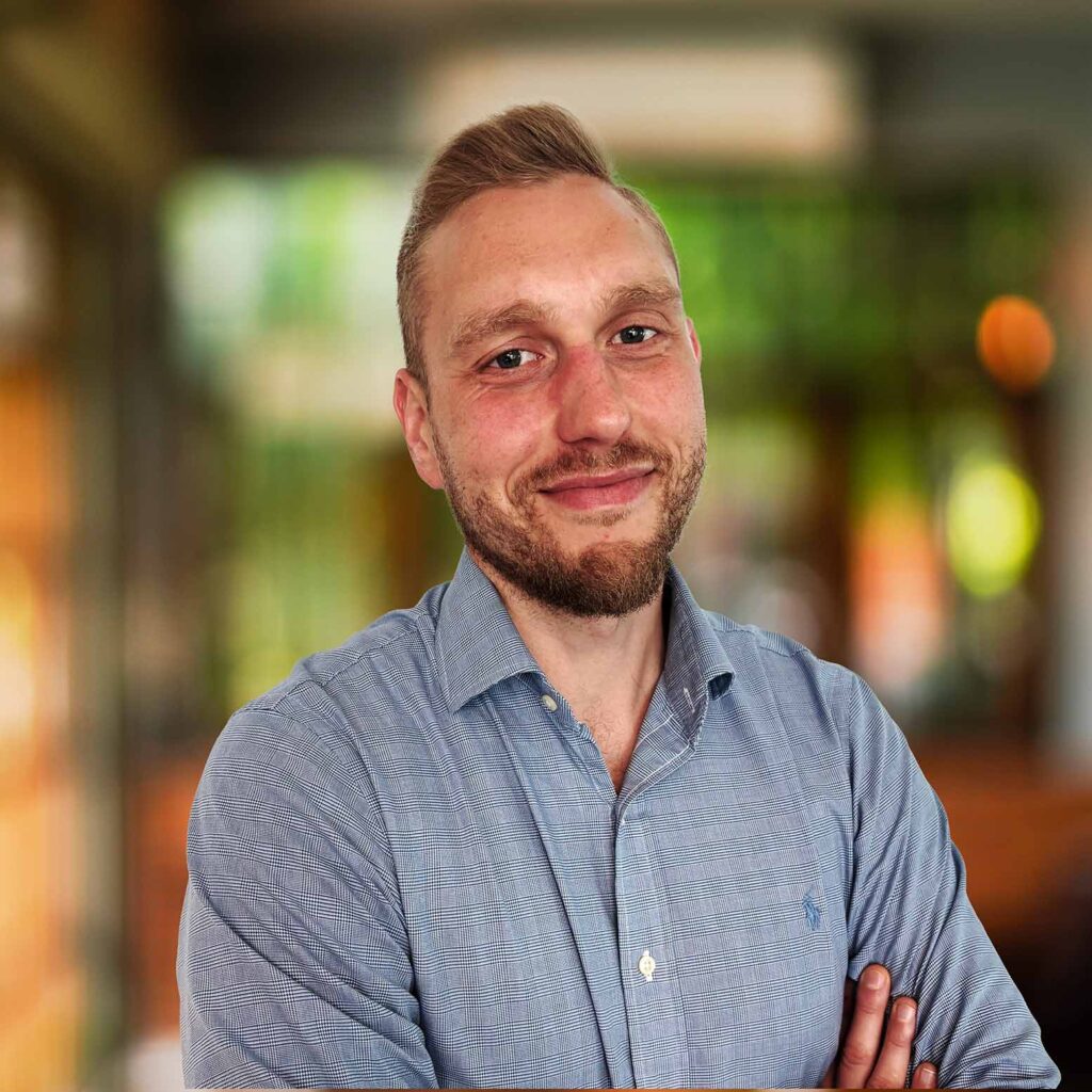 The image shows a man with a friendly expression, standing with his arms crossed. He has short, light brown hair styled upward, a trimmed beard, and a slightly flushed face. He is wearing a light blue checkered button-up shirt. The background is softly blurred, suggesting an indoor setting with warm lighting, possibly in a modern office or a casual professional environment. The overall mood is approachable and confident.