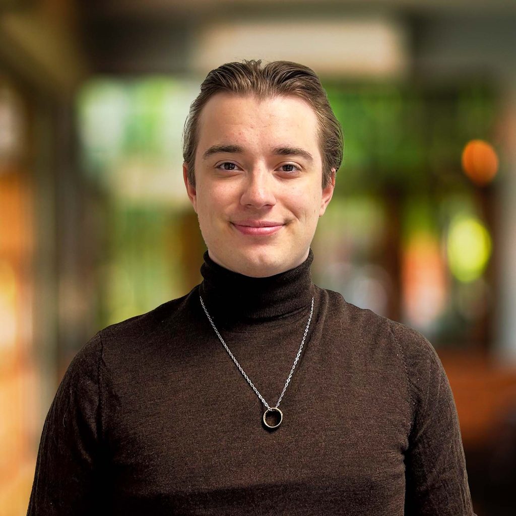 The image shows a young man with short, slicked-back brown hair, wearing a dark brown turtleneck sweater. He has a gentle smile and is wearing a silver necklace with a circular pendant. The background is softly blurred, displaying a warm and vibrant indoor environment with green, yellow, and orange hues, suggesting a cozy, well-lit setting.